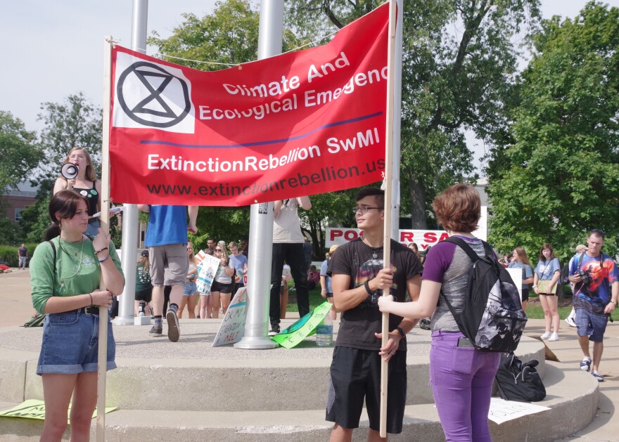 Three young people hold a bright red banner that says "climate and ecological emergency - extinction rebellion SwMI - www.extinctionrebellion.us"