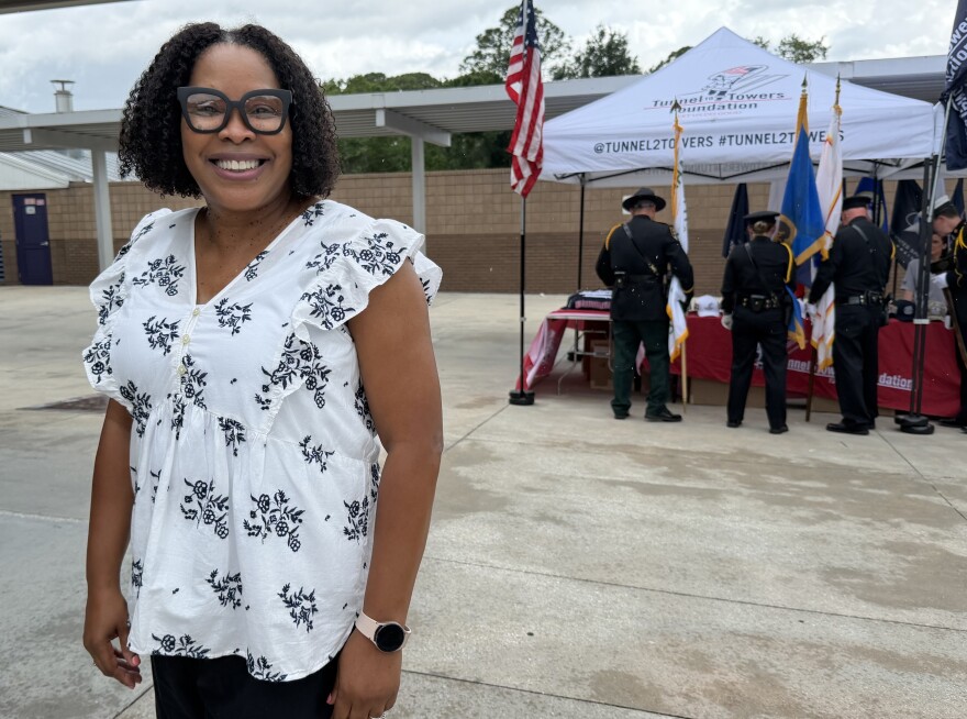 A woman with large black glasses and a white short with black flowers stands near a Tunnel to Towers tent at Booker Elementary
