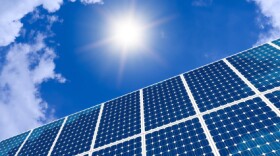 A series of solar panels outlined against a bright blue sky, with clouds. The sun shines down on the solar panels in this solar farm.