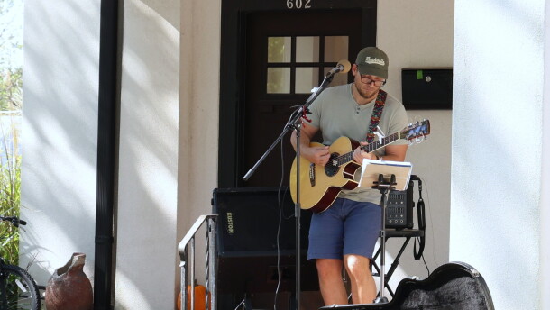Trevor C. Smith performs at the 10th annual North Heights Porchfest in Joplin on Oct. 10, 2026. He's on a front porch, standing behind a microphone and in front of a speaker. He's playing an acoustic guitar with the case open in front of him.