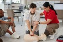 A woman coaches a man doing CPR chest compressions on a mannequin.