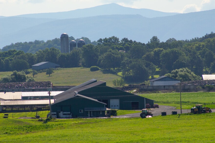 English: A photo of large farm buildings set into green fields, with a blue-colored hill in the background. Español: Una foto de grandes edificios agrícolas sobre campos verdes, con una colina de tono azul en el fondo.