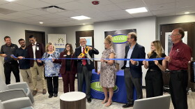 A man holding a large pair of ceremonial scissors cuts a ribbon held by multiple people to open a new building. 