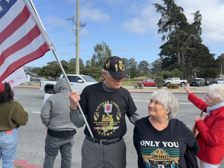 An older couple look at each other on the sidewalk at a protest. The man is holding an American flag and wearing a veteran t-shirt and cap.