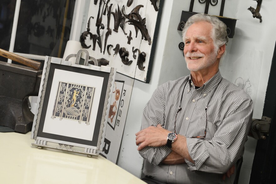 A man smiles while seated next to framed artwork