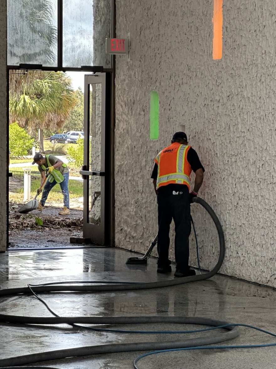 A person in a high visibility vest vaccums water inside a building with the doors open where you can see a man outside shoveling tree limbs.