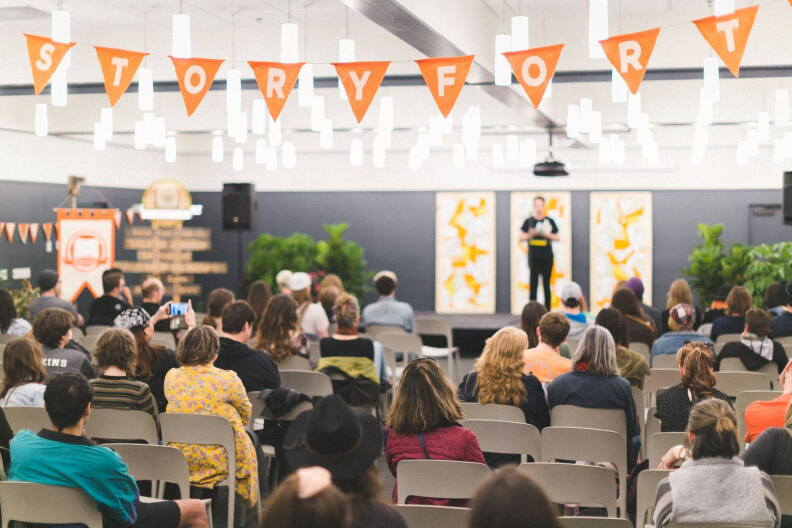 Crowd attends Storyfort with orange flags spelling out Storyfort in white hanging above. 