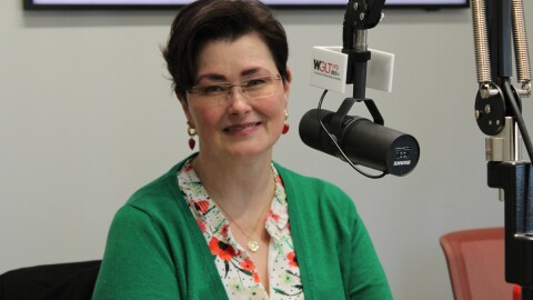 A woman in a green blouse and eyeglasses sits at a desk with a microphone in front of her