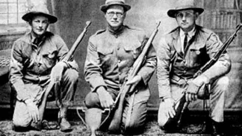 Three World War I era soldiers or rifle team members kneeling in uniform, holding bolt-action rifles, with a trophy placed on the floor in front of them.