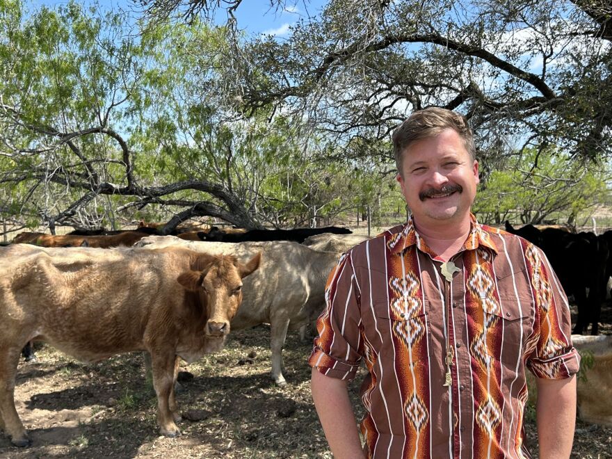 Max Kruemke, co-owner of Bastrop Cattle Company, stands in a shady part of a pasture next to some of his cows. Kruemke is wearing a maroon and white patterned short sleeve shirt, and cow head bolo tie.