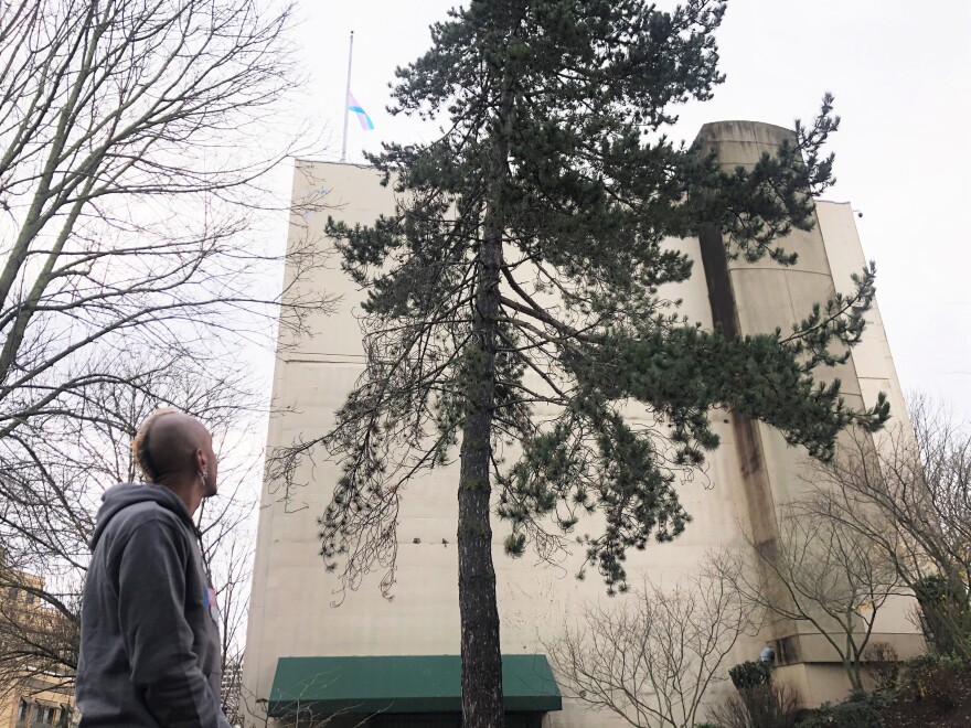 A slender man with a short blonde mohawk haircut looks up to at a pink, white, and blue trans flag at half mast on top of building.