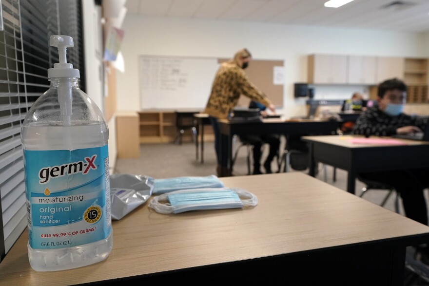 Hand sanitizer, wipes, and surgical masks rest on a desk in a classroom.