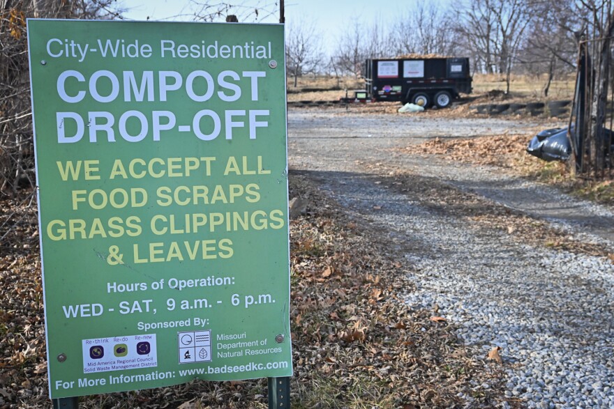 A large green sign sits on the ground next to a gravel road. In the background a dumpster sits with a pile of brown leaves piling up inside. The sign reads "City-Wide Residential Compost Drop-Off, we accept all food scraps, grass clippings and leaves."