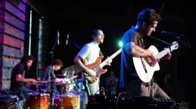  A man plays acoustic guitar while standing near a microphone, with a man playing bass and two other men playing drums behind him.