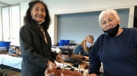 Two women stand together near a table with handwoven baskets.