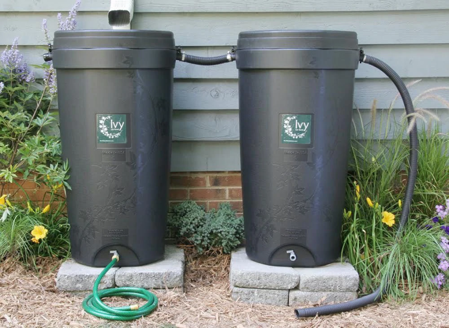 Two black barrels sit on concrete slabs against a grey house amidst flowers.