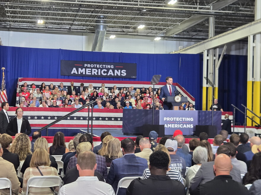 Vice President JD Vance speaks at a podium before a large audience seated in an industrial venue. Red, white, and blue decorations surround the stage, with signs reading “Protecting Americans.” A teleprompter stands beside Vance, and security personnel and supporters are visible in the scene.