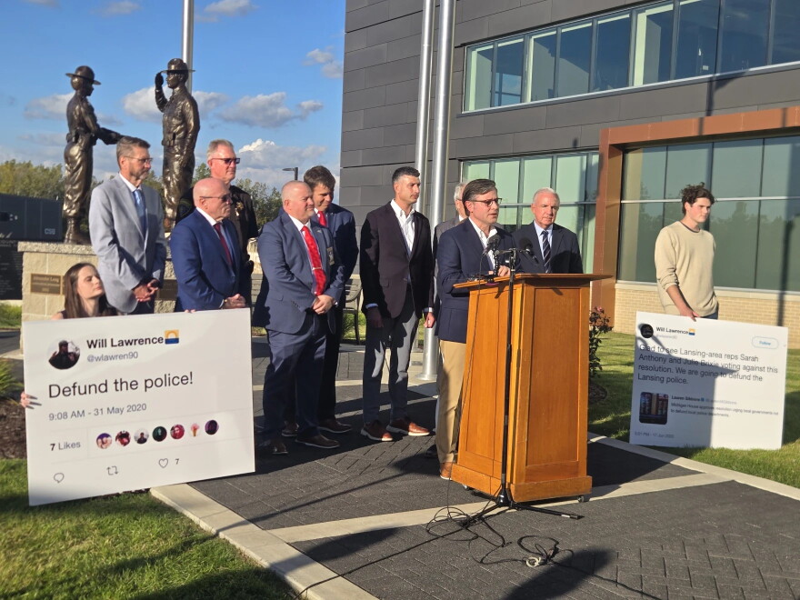 U.S. House Speaker Mike Johnson lends his support to the reelection campaign of Congressman Tom Barrett (R-MI 7) on October 7, 2026. Behind Johnson, Barrett supporters hold posters showing deleted tweets by Barrett's Democratic opponent Will Lawrence.