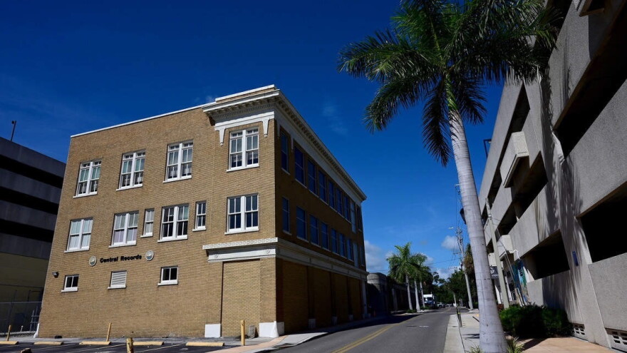 A tan, three-story building stands on the left, next to a road lined with palm trees, a parking garage on the other side of the street.