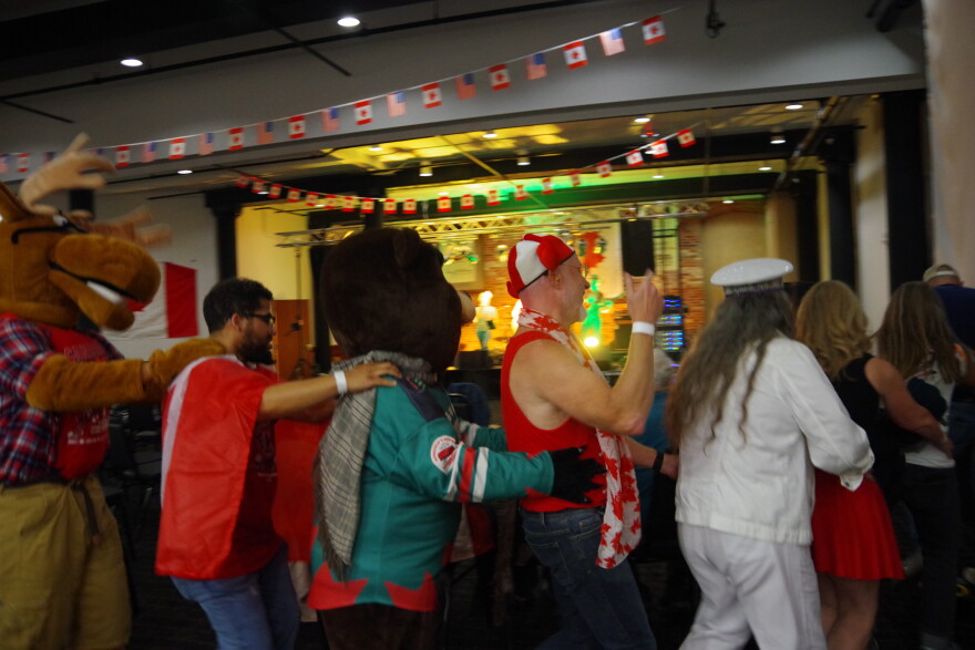 A line of people are pictured in a room where the ceiling is decorated with dozens of tiny american and canadian flags. The people have their hands on eachother's shoulders and hips in a conga line. In the line is a moose mascot wearing a plaid flannel, a man draped in a canadian flag, a beaver mascot wearing a hockey jersey, a man in a canadian flag hat with red moose ears, and a woman in a white jean jacket and white jean pants wearing a white conductor's hat. 