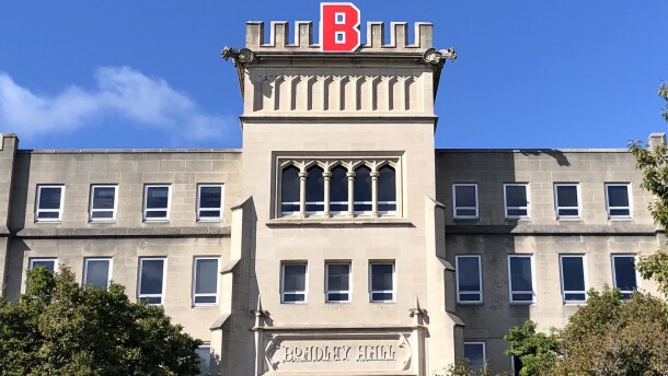 A stone academic building with a “B” in red on top, reading “Bradley Hall” above the entrance, with trees and a clear blue sky.
