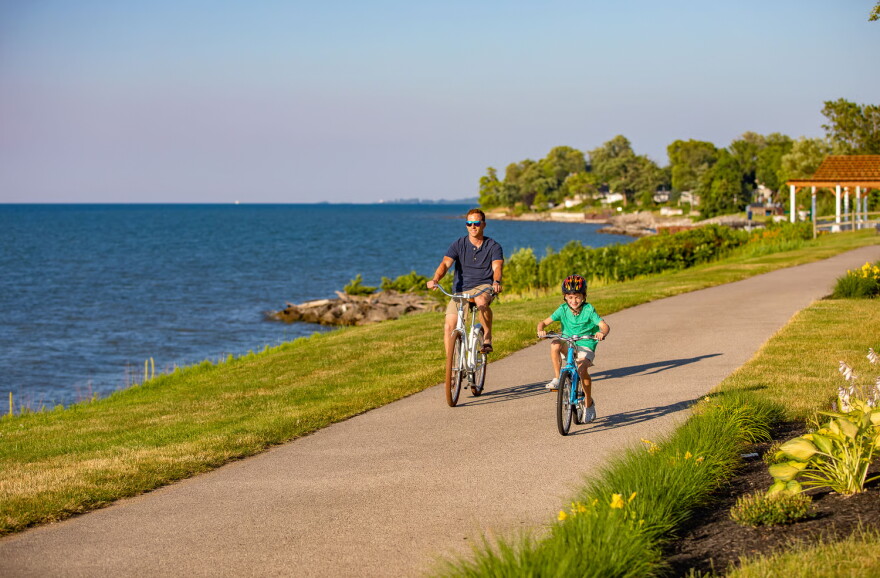 Man and boy ride bicycles down paved path by water.