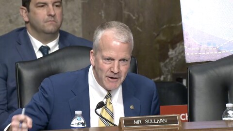 a man in a suit and tie pounds the table in a hearing room. A nameplate identifies him as "Mr. Sullivan."