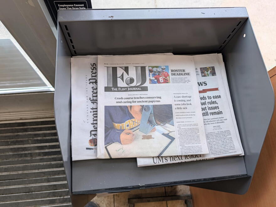 The Flint Journal sits on top of a copy of The Detroit News and the Detroit Free Press in a grey, metal newsstand