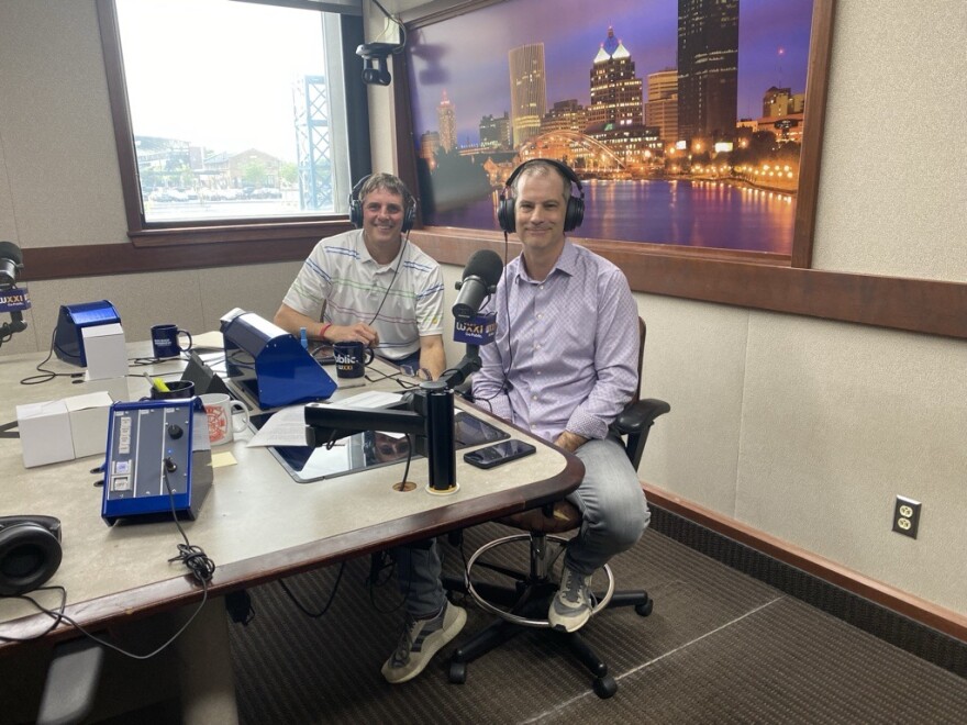 Two smiling men wearing headphones sit at a table in a radio talk studio: A man at left has short light hair and is wearing a patterned polo shirt; a man at right has short dark hair and is wearing a light purple button-down shirt