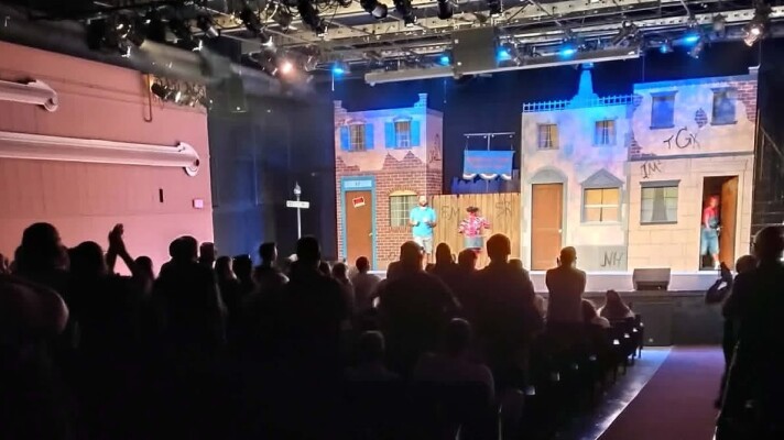 A crowd of people at a theater standing up clapping for a stage with three people and a fake street behind them.