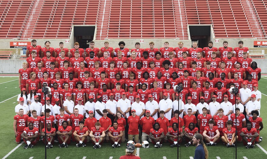 Eight long rows of college football players, coaches and support staff posing for a photo in a football stadium. The players are all wearing red jerseys with while numbers and 'Redbirds' at the top of the jerseys. 