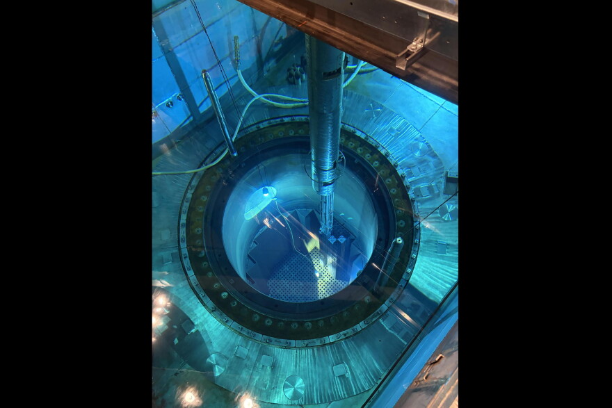 The image is shot from above a pool of blue water. A circular hole sits inside, just below other parts of the submerged metal. A silver metal rod can be seen above the water holding a smaller metal rod that is partially submerged. Various pipes and tubes also feed in and out of the pool. 