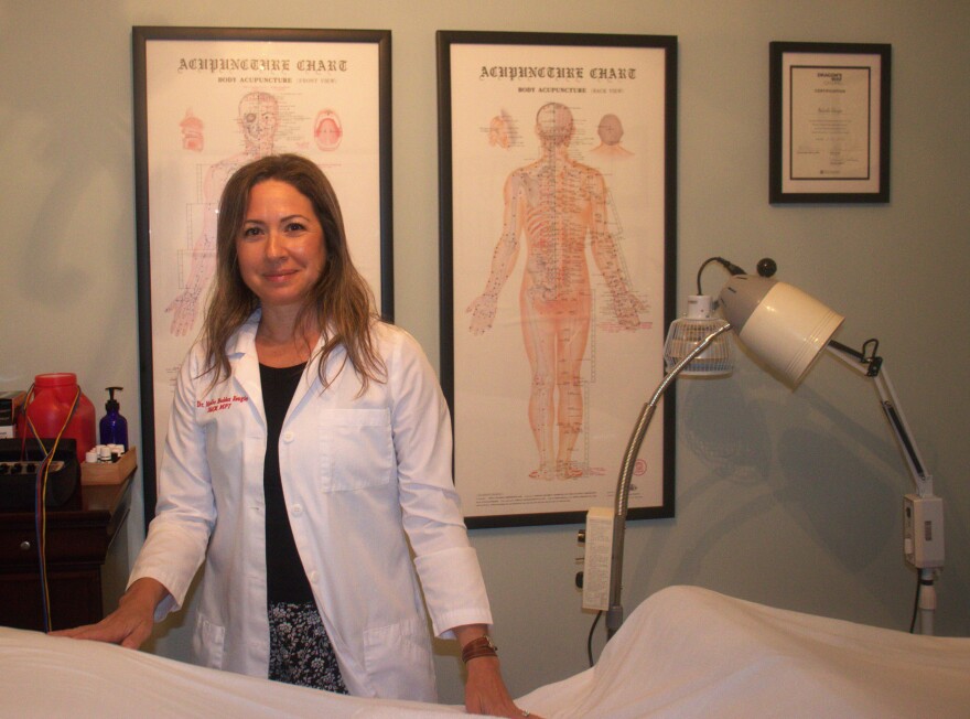 A blonde woman in a white lab coat stands behind two diagrams of th human body and a bed draped with a white sheet.