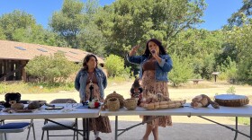 Two women wearing matching outfits stand in front a tables full of artifacts with green shrubs behind them. 