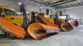 A dump truck sits in the foreground of a large garage, with a yellow cab that has two orange snowplow attachments, while several identical trucks recede into the background.