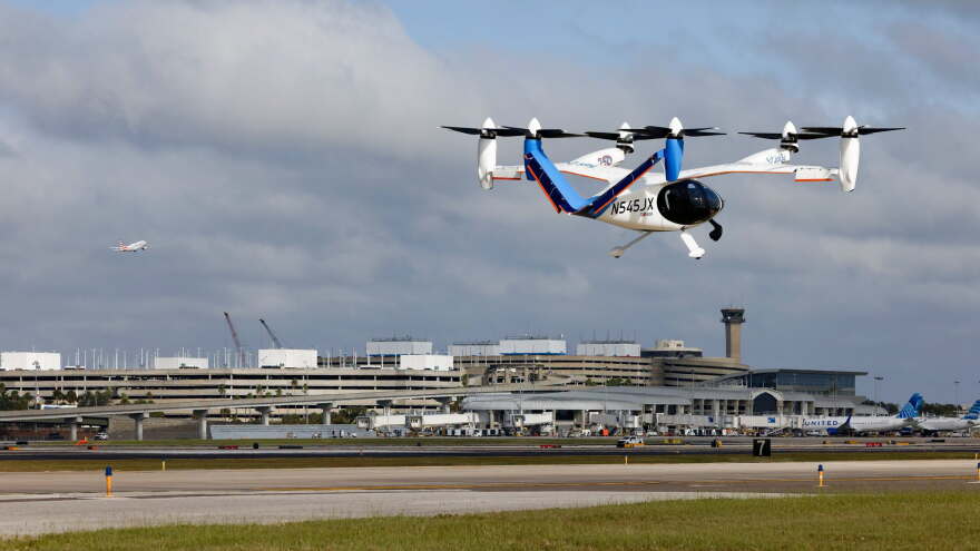 A white and blue electric aircraft flies at an airport during a demonstration flight, with six propellers visible.