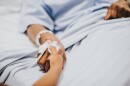 Hospital patient lies in a bed with an IV hooked up to their hand. Child holds the patient's hand.