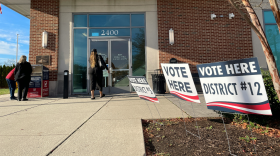 Photo of the outside of a  town hall. There are several signs saying "vote here" for different districts. One person is walking inside while two people are standing near a ballot drop box.