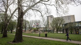 A large green space shows many trees  with a person walking on a brick pathway. A large, multistory hotel can be seen in the background.