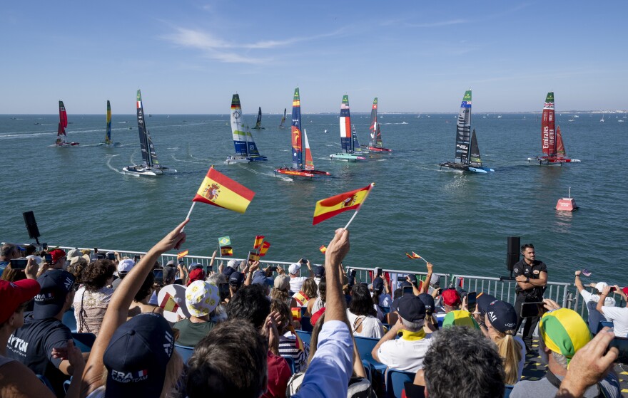 Spectators wave their flags as they watch the action from the grandstand in the race stadium as Emirates Great Britain SailGP Team helmed by Dylan Fletcher led ahead of New Zealand SailGP Team helmed by Peter Burling, Spain SailGP Team helmed by Diego Botin and the fleet on Race Day 1 of the DP World Spain Sail Grand Prix held in Cadiz, Spain Saturday 4 October 2025. Rolex SailGP Championship Event 11 2025 Season. Photo: Andrew Baker for SailGP. Handout image supplied by SailGP