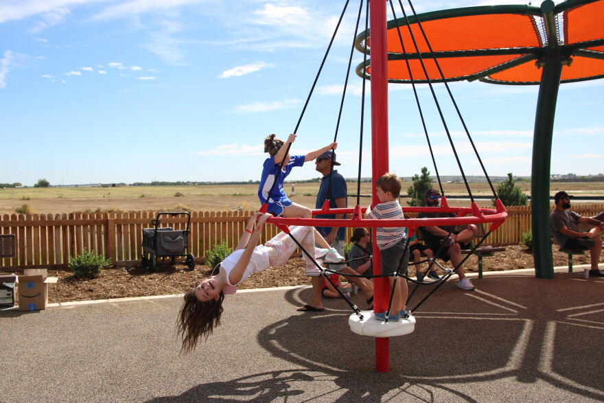 A young girl hangs on a metal bar and leans back, her hair falling down to the ground. She is smiling. She and other kids are hanging onto a red playground structure with black ropes that spins around. There's an orange shade structure behind them.