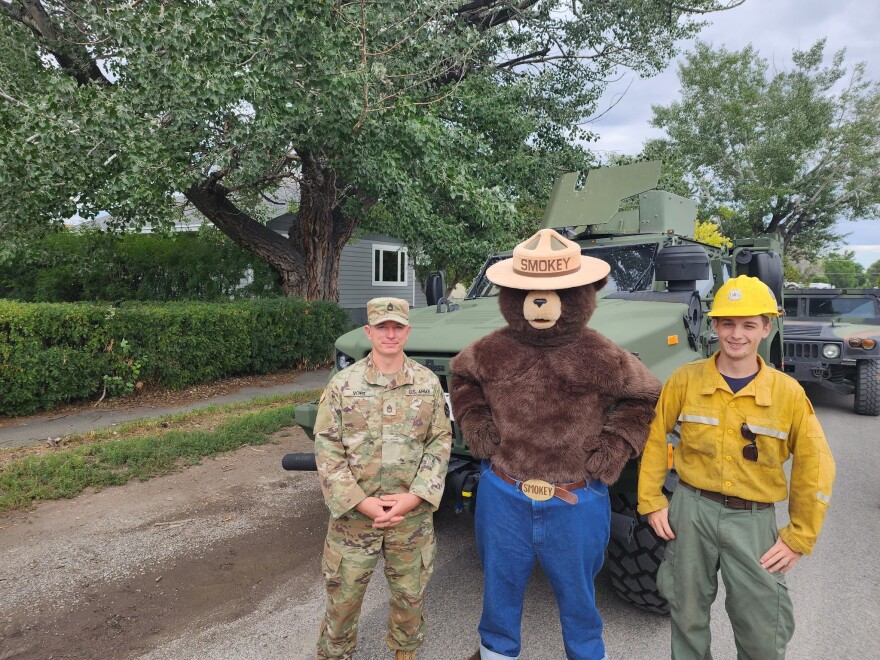 A person in a camouflage uniform stands next to someone in a Smokey Bear costume and another person in a yellow long-sleeve shirt and yellow hard hat. Behind them is a Corps vehicle, a tree and a green house.