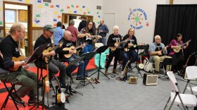 Line of eight people in a semicircle playing ukeleles with a row of people standing behind them with a sign painted on the wall that reads 'he's got the whole world in his hands'