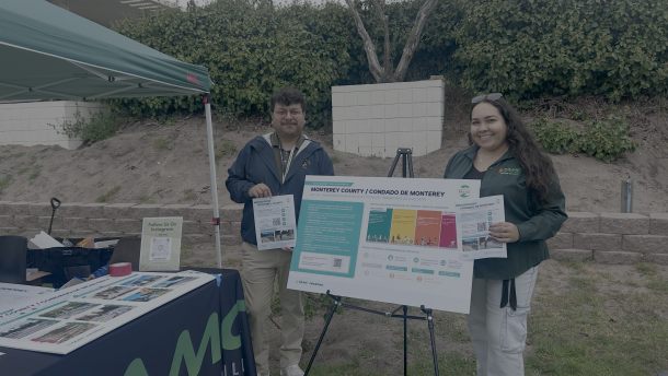 Two people standing holding flyers with a poster board on a stand between them. They are standing outside, a booth with a pop-up tent is to their left.