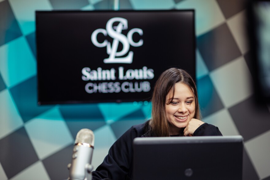 A smiling Thalia Cervantes Landeiro sits at computer and a microphone. A Saint Louis Chess Club sign is in the background.