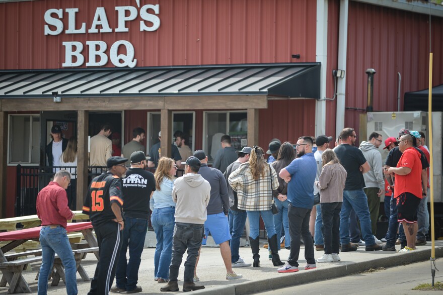 A long ling of people stand outside a red, metal building. There are picnic tables outside and the sign on the building reads "Slap's BBQ."