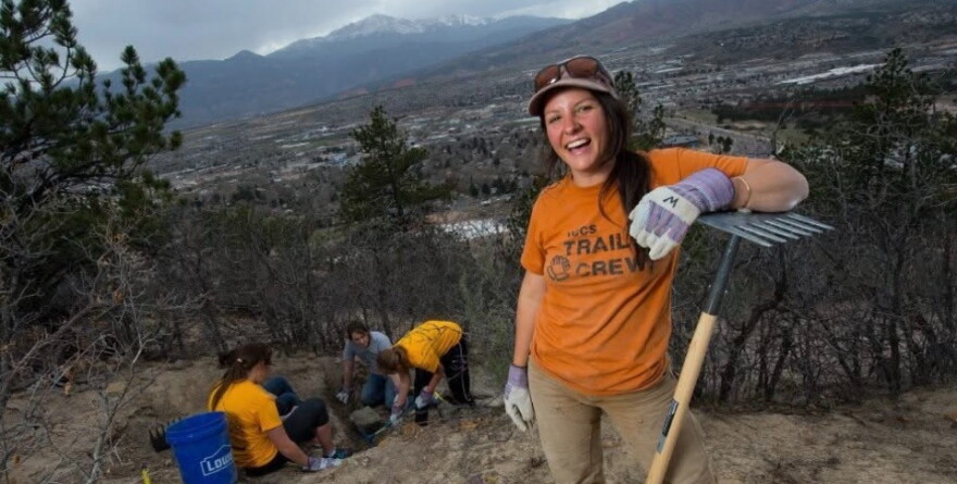 A woman in an orange shirt leans against a rake in front of people digging a hole in the dirt.