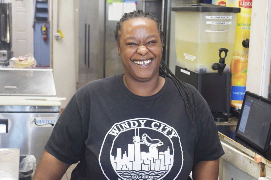 Woman smiling and standing next to a restaurant kitchen and wearing a black shirt with a skyline logo and "Windy City" inscribed on the front.