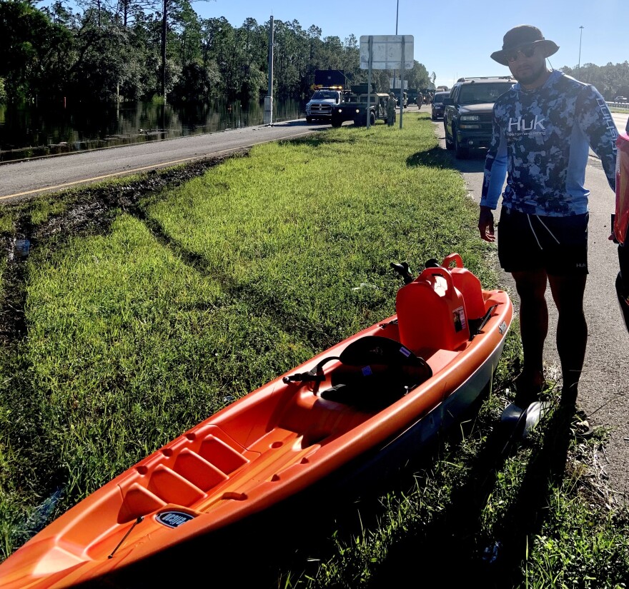 Man in shorts, blue shirt and sunhat stands next to orange kayak.