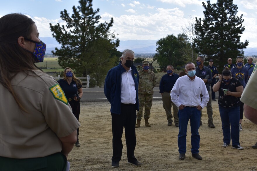 Sisolak and Brooks are both wearing white button up shirts and masks. They are standing outside on dirt, surrounded by National Guard and firefighting personnel.